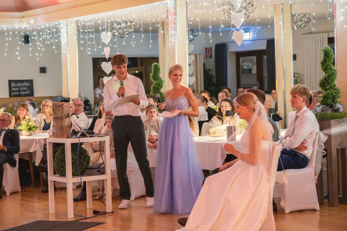 Rede im Festsaal unter Lichterketten, die Braut hört lachend zu.