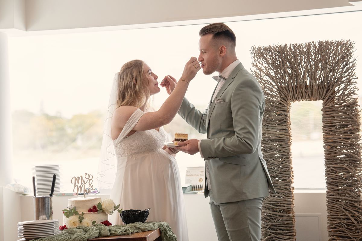 Braut füttert den Bräutigam mit Hochzeitstorte vor einem hellen Fenster.