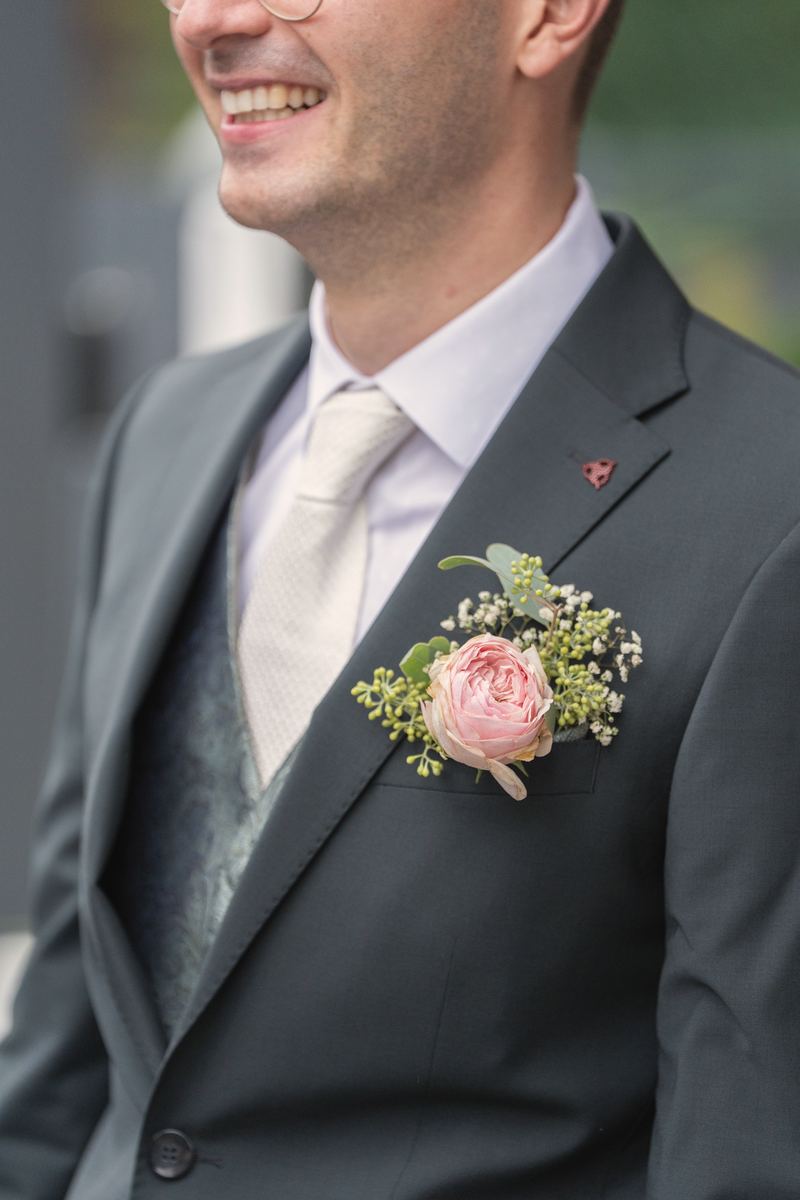 Lachender Bräutigam im dunklen Anzug mit Rosen-Anstecker am Revers.